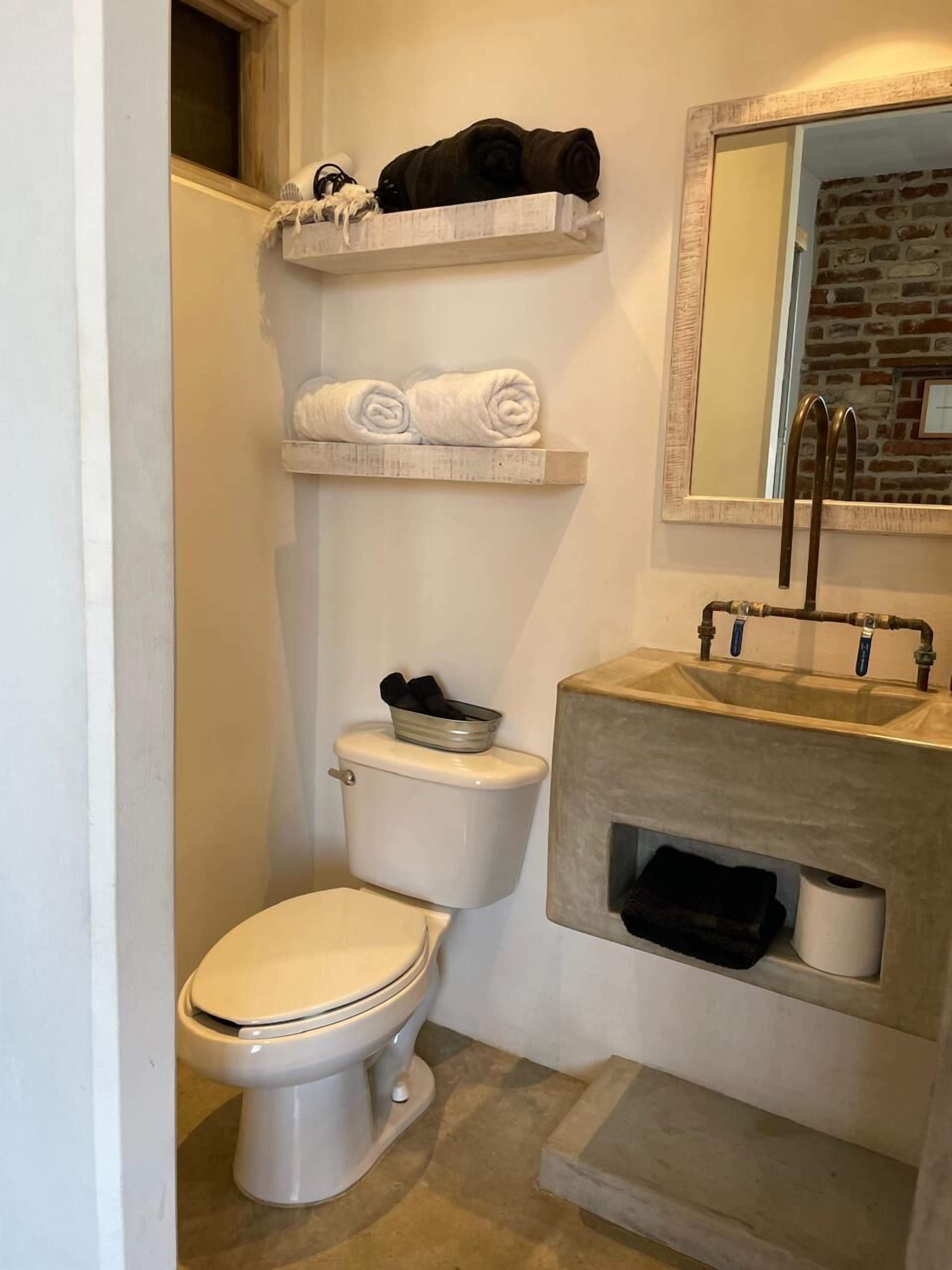 Une salle de bain avec des toilettes, un lavabo et un miroir. On y trouve des serviettes et un panier sur une étagère située au-dessus des toilettes.