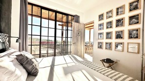 A bedroom with a large window, a bed with striped bedding, a nightstand, and a wall with framed pictures. - Baja House (Todos Santos)