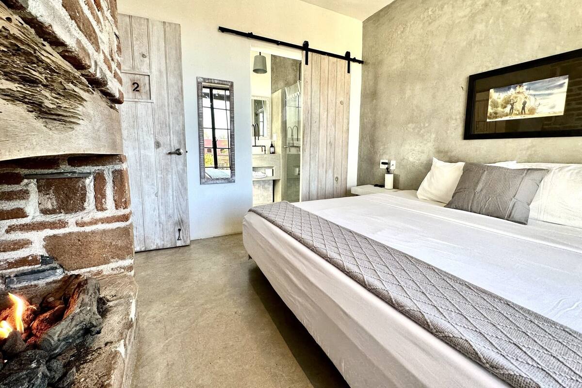 Ein Schlafzimmer mit Kamin aus Backsteinen, einem Bett mit gesteppter Bettdecke, einem Wandbild und Blick durch ein Fenster.