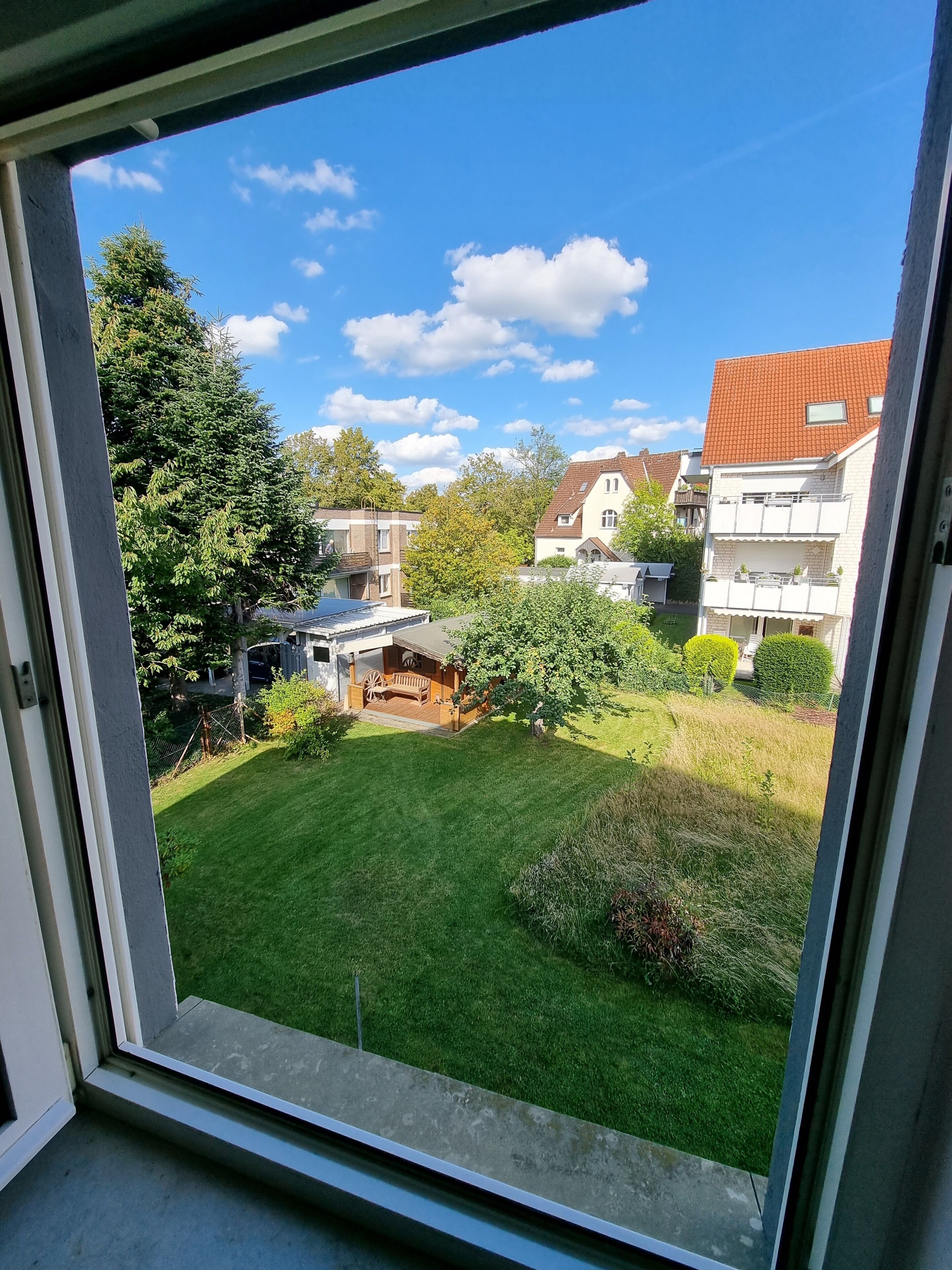 Blick durch ein Fenster auf eine Wohngegend mit Rasen, einer Holzterrasse und Häusern mit roten Dächern.