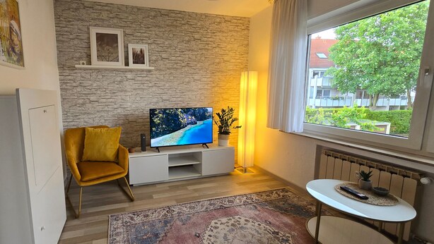 Una sala de estar moderna con una pared de piedra, un sofá, un televisor, una mesita y vistas a una zona residencial.