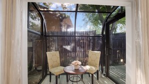Une véranda avec une table ronde et deux chaises, un vase rempli de fleurs et une vue sur une clôture en bois et des arbres.
