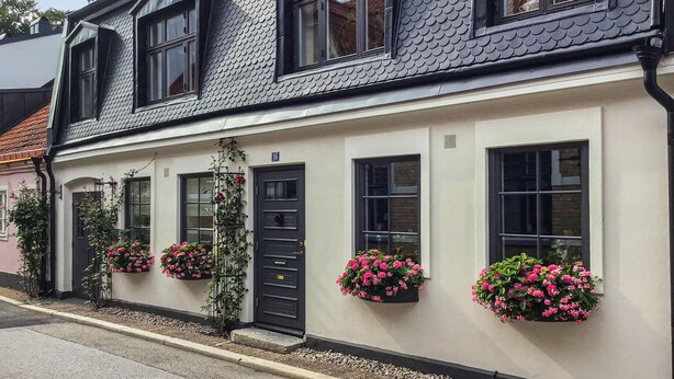 A two-story building with a dark roof and white walls. There are flower boxes with pink flowers on the windowsills. - 2 bedroom nice home in Ystad (Ystad)