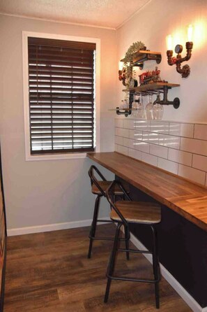 A kitchen with a wooden countertop, two bar stools, a window with blinds, and wall-mounted shelves with decorative items. - Cozy & Convenient Bungalow - Downtown & HPU (Brownwood)