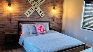 A bedroom with a brick wall, a bed with a striped bedspread, two bedside lamps, and a crossroad sign on the wall. - Cozy & Convenient Bungalow - Downtown & HPU (Brownwood)