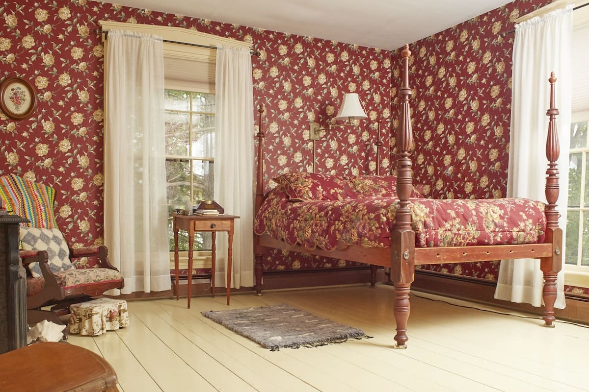 A room with a four-poster bed, a rocking chair, a small table, and a window with curtains.