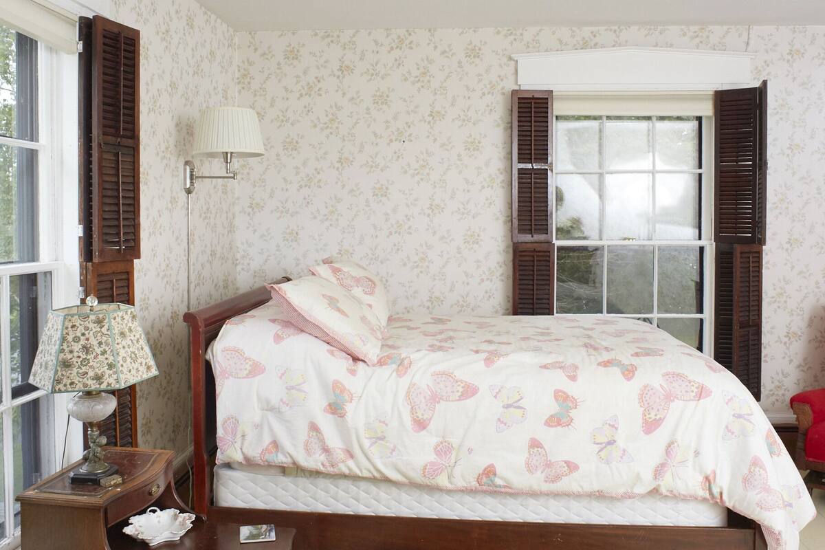 A bedroom with a bed, a bedside table, a lamp, a window with shutters, and a patterned rug.
