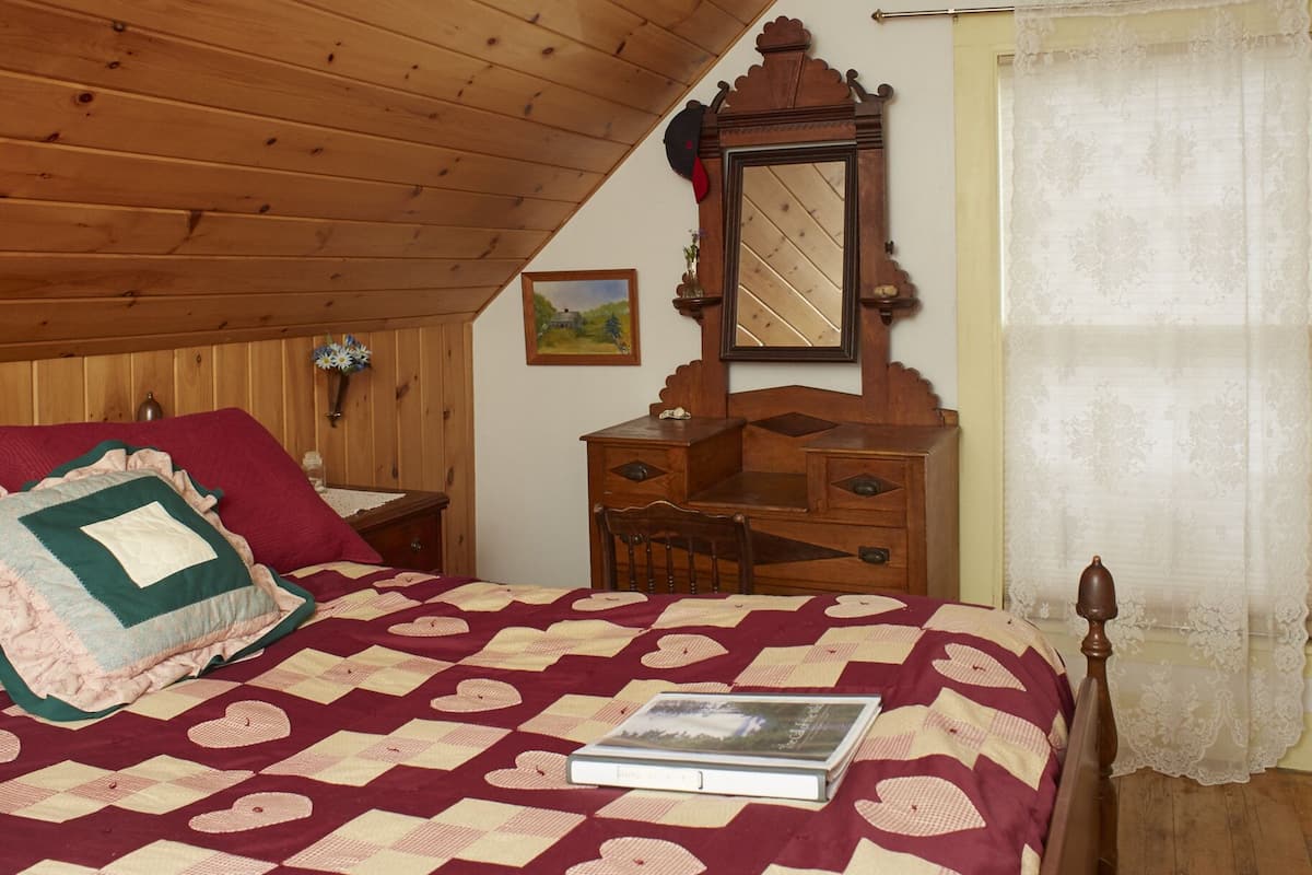 A bedroom with a wooden ceiling, a bed with a quilt, a dresser, a mirror, and a window with curtains.