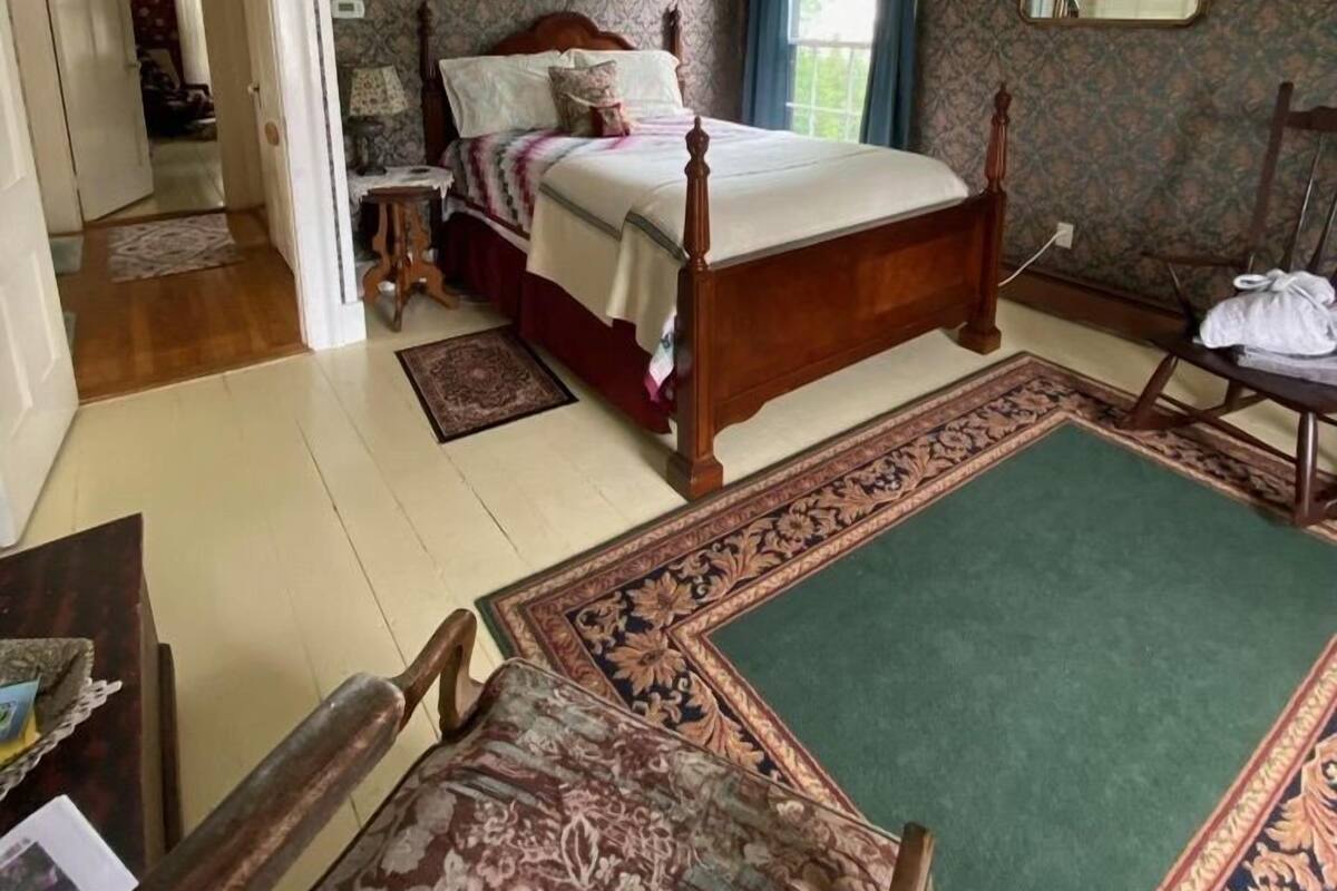 A bedroom with a wooden four-poster bed, a green area rug, a patterned carpet, a wooden chair, and a mirror on the wall.