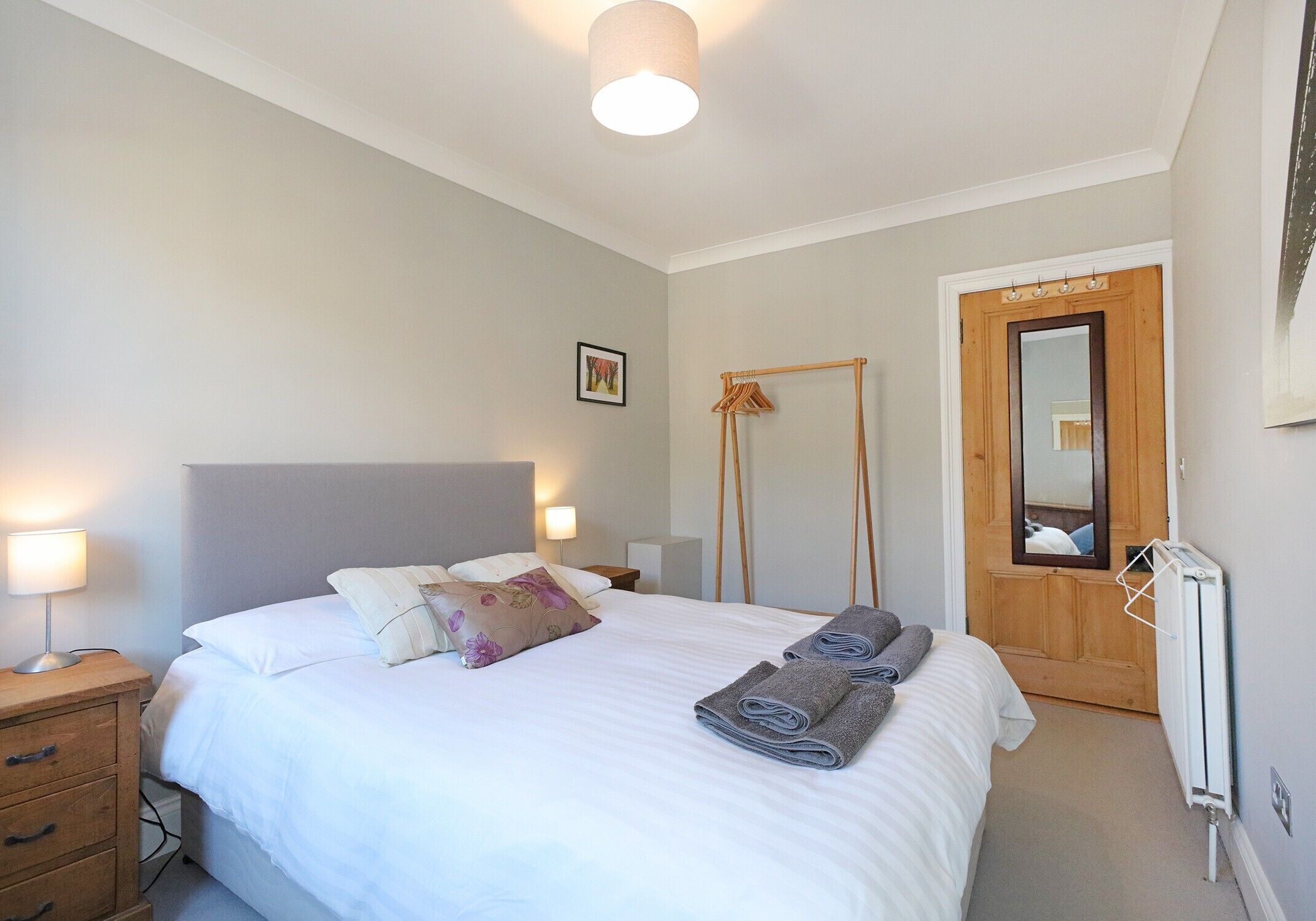 En pænt redt seng med hvide sengetøj og to sammenrullede håndklæder, et natbord med lampe, en træskab og et spejl på døren.