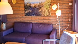 Una habitación con pared de ladrillo, un sofá morado, una mesa de madera, una lámpara y un cuadro con una playa.