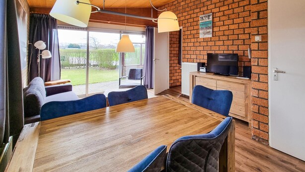 Una habitación moderna con una mesa de comedor de madera, sillas azules, un televisor y una pared de ladrillo.