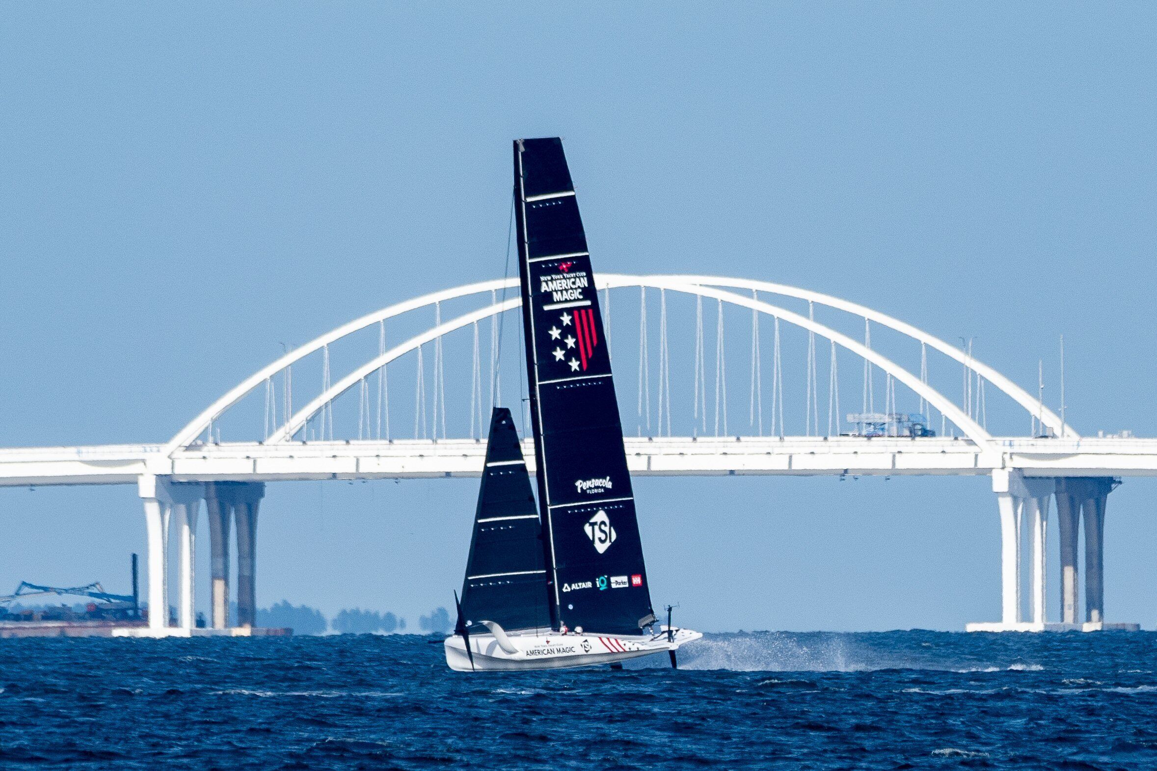A sailboat with a large sail and a logo on the back, sailing under a bridge.