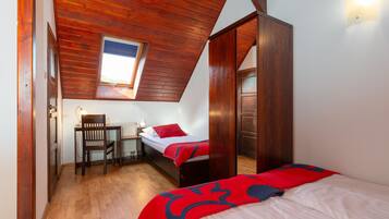 A bedroom with a sloped ceiling, a wooden desk, a chair, a bed with a red and blue blanket, and a window with daylight.