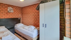 Een kamer met een bakstenen muur, een bed met wit beddengoed, een kast met blauwe handgrepen en een klein plankje met spullen.