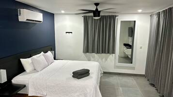 Ein Hotelzimmer mit Bett, Deckenventilator, Klimaanlage, Spiegel und grauen Vorhängen.