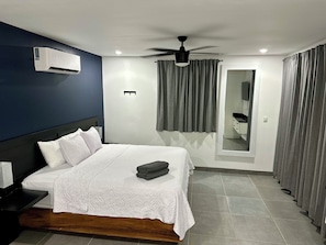 A hotel room with a bed, a ceiling fan, an air conditioning unit, a mirror, and grey curtains.