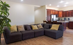 A living room with a sectional sofa, wooden flooring, and a kitchen area with wooden cabinets and appliances.