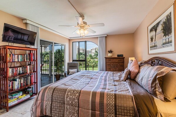 Ein Schlafzimmer mit einem großen Bett, einem Deckenventilator, einem Fernseher, einem Bücherregal und Blick auf Palmen durch das Fenster.