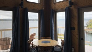 A wooden cabin with a round table and chairs, large windows, and a view of a body of water.