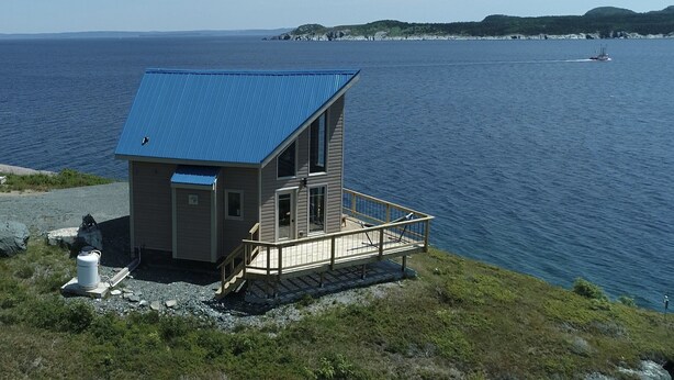 A small, single-story cabin with a blue roof, situated on a rocky shore with a view of the sea and distant hills.