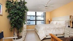 Une chambre à coucher comprenant un lit, des tables de chevet, un ventilateur de plafond, une fenêtre donnant sur des immeubles et une plante en pot.