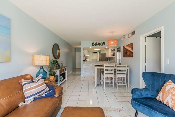 Uma sala de estar com um sofá marrom, uma poltrona azul e um espaço de jantar, com uma cozinha ao fundo.