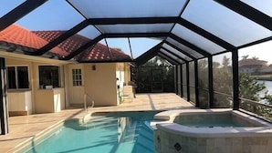 Une piscine avec un jacuzzi, entourée d’un patio couvert offrant une vue sur une maison et un plan d’eau.