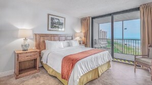 Habitación de hotel con cama, mesita de noche, silla y balcón con vista a la playa y edificios.