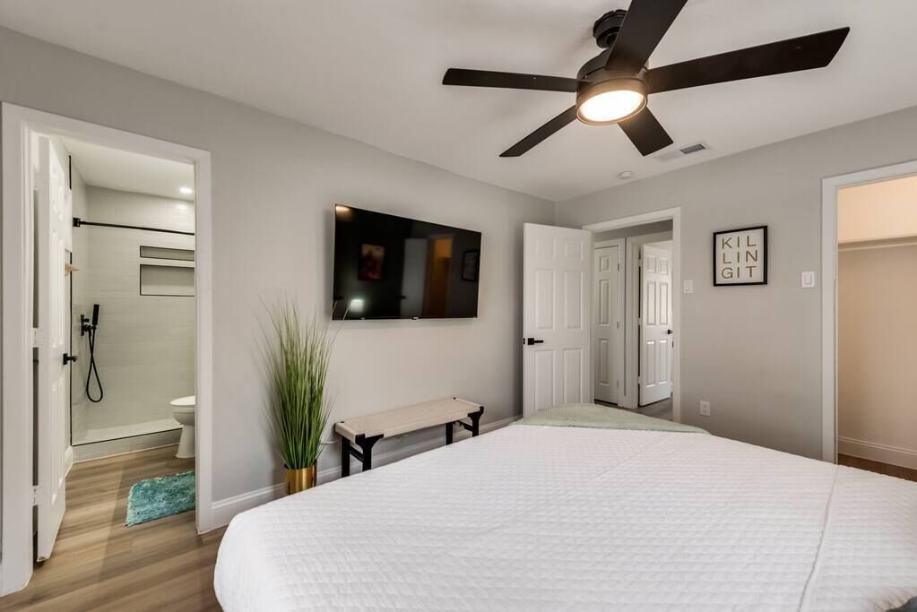 Un dormitorio con cama, ventilador de techo, televisor y puerta que da a un baño.