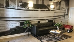 Un salón con un gran mural de un auditorio, un sofá de cuero negro, una mesita de centro y un carrito de juguete rojo.