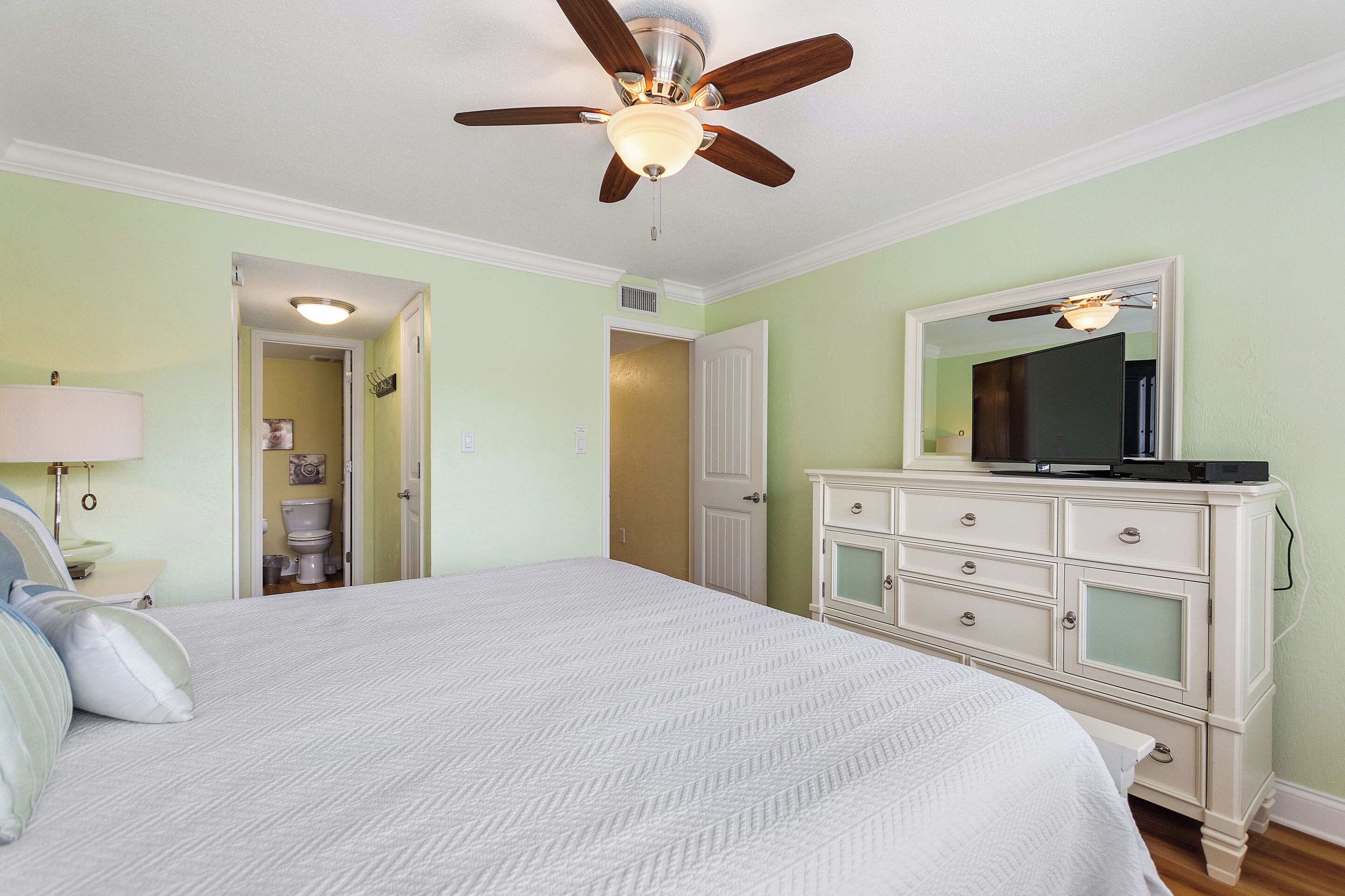 Un dormitorio con cama, cómoda, ventilador de techo y un baño visible a través de una puerta abierta.