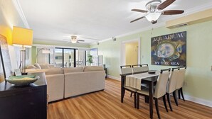 A spacious living room with a wooden floor, a dining area with a table and chairs, a ceiling fan, and a large aquarium painting on the wall.