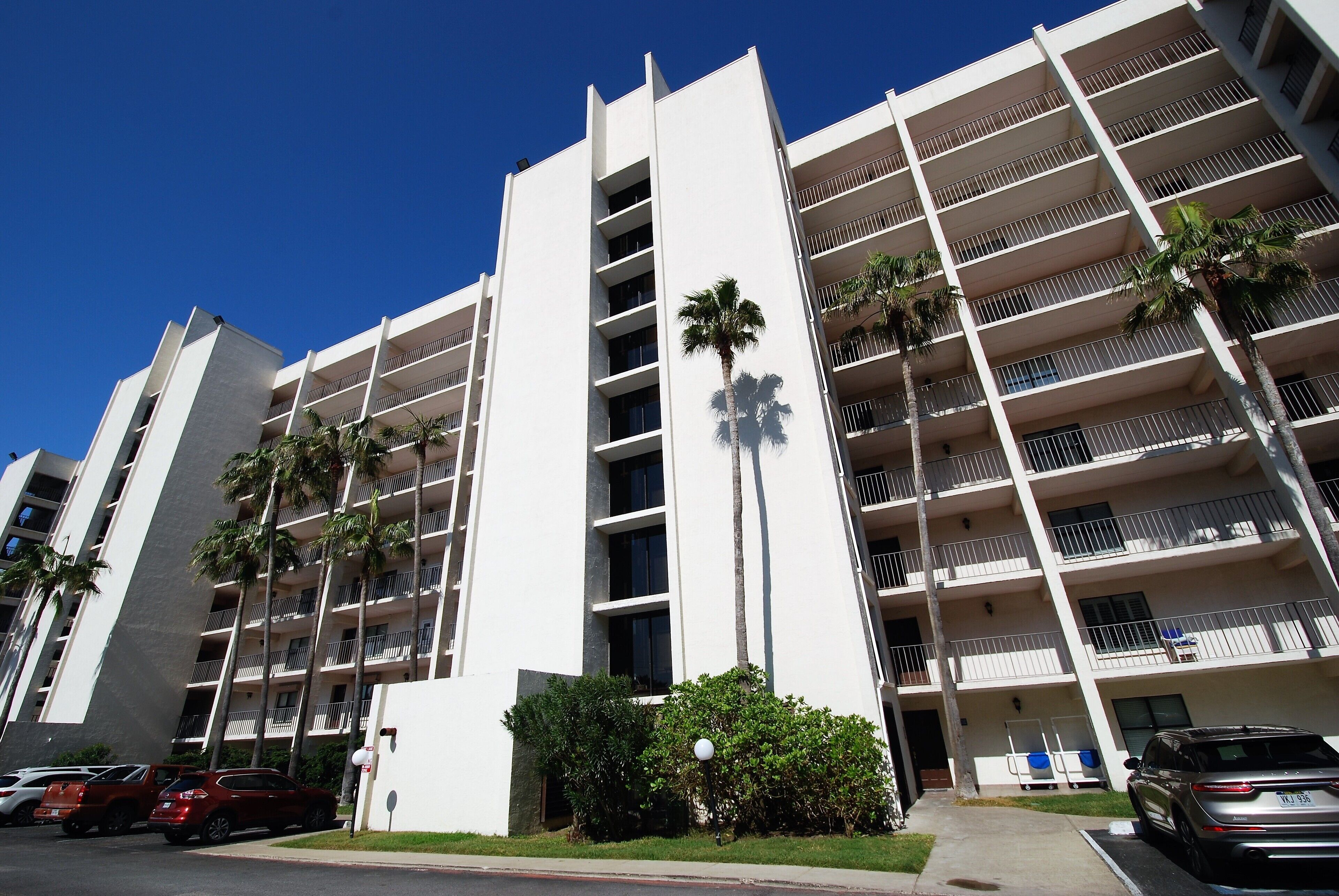 A multi-story building with balconies, palm trees, and parked cars.