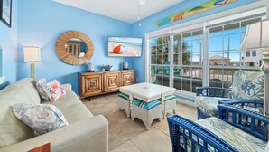A room with a white sofa, blue chairs, a wooden cabinet, a round mirror, a television, and a large window with a view of palm trees. - MY FRIEND'S PLACE EAST - COSTAL COTTAGE, GULF VIEWS FROM DECK, SUPER COZY (Gulf Shores)