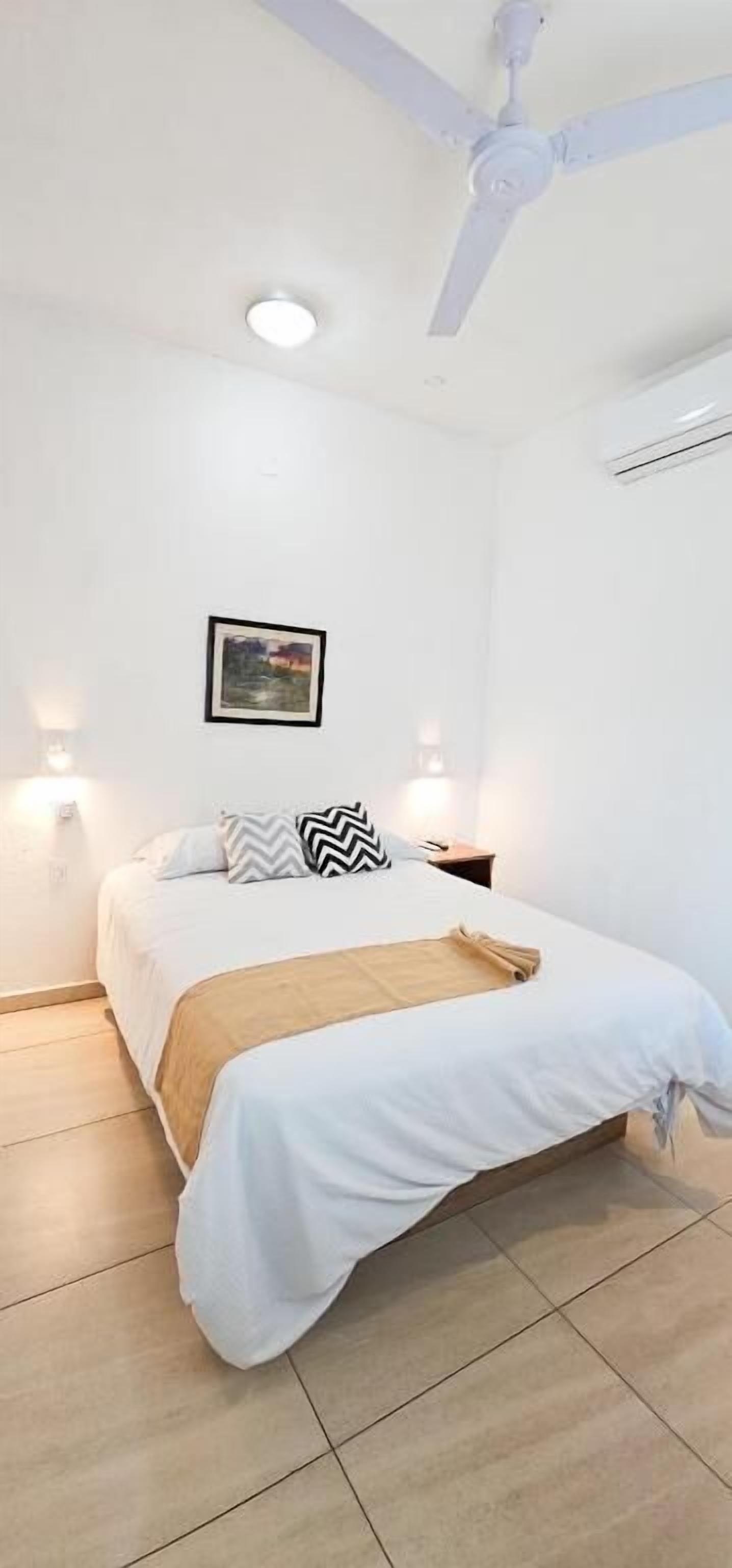 A bedroom with a bed, white walls, a ceiling fan, two bedside lamps, and a framed picture on the wall.