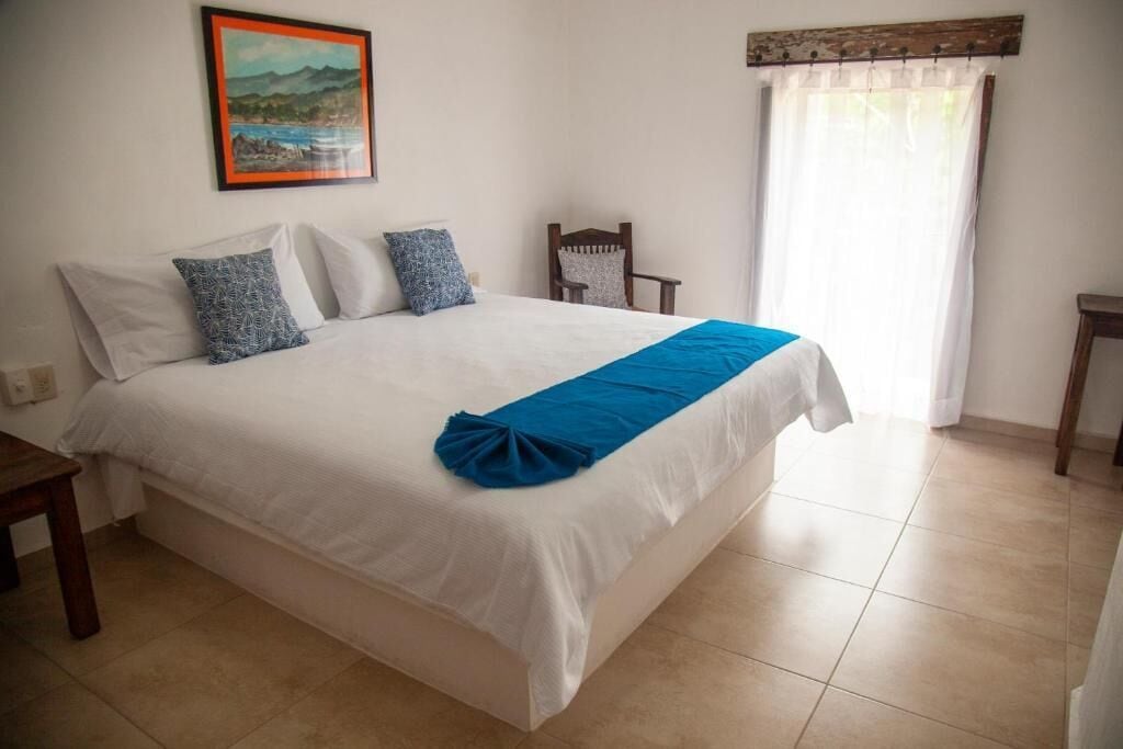 Un dormitorio con cama, almohadas, una silla, una mesita y un cuadro en la pared.
