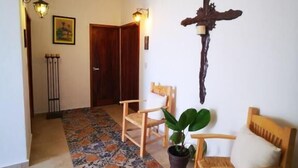A room with a patterned floor, wooden chairs, a potted plant, a wall-mounted cross, and a door in the background. - La Familia Hotel y Restaurante   (San Blas)