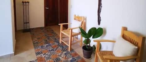 A room with a patterned floor, wooden chairs, a potted plant, a wall-mounted cross, and a door in the background.