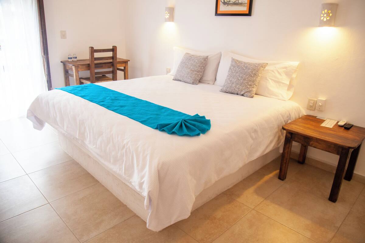 Una cama bien tendida con un detalle azul, una mesita de noche de madera con un teléfono y una silla en una habitación con paredes blancas y suelo de baldosas.