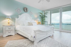 Ein Schlafzimmer mit einem weißen Bett, einem Nachttisch mit Lampe, einem großen Fenster mit Meerblick und einer Zimmerpflanze.