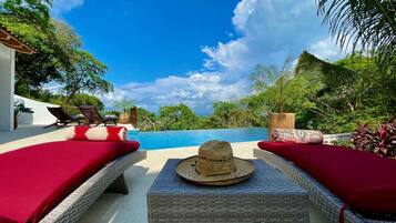 A poolside area with wicker lounge chairs and a table, a hat, and a view of the sky and trees.