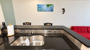 A kitchen with a stainless steel sink, black countertop, and a red couch in the background.