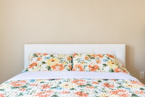 Een bed met een deken en kussens met een bloemenprint, een wit hoofdbord en een effen muur.