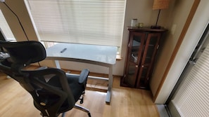 Een kamer met een houten vloer, een zwarte bureaustoel, een tafeltje met een afstandsbediening en een kast met een lamp.