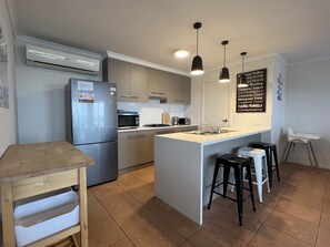 A modern kitchen with a central island, a refrigerator, microwave, and a sink. There are three bar stools and a high chair. - The Sandcastle, KI - Esplanade location (Kingscote)