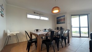Un comedor con mesa de madera, sillas negras, una trona, una lámpara en el techo, una ventana y una puerta que da al exterior.