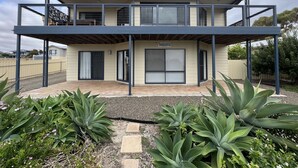 Una casa de dos pisos con un porche cubierto, un letrero que dice "SANDCASTLE" y un patio trasero cercado con diversas plantas.