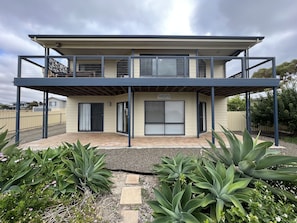 A two-story house with a covered patio area, a sign reading "SANDCASTLE," and a fenced backyard with various plants. - The Sandcastle, KI - Esplanade location (Kingscote)