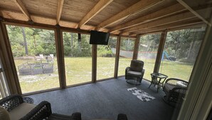 Une terrasse couverte, aménagée avec des meubles en osier, un téléviseur et offrant une vue sur une pelouse.